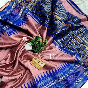 May include: A woven textile featuring a dusty rose center with a prominent blue border. The border displays intricate ikat patterns in black, white, and blue, accented by a sharp triangular motif. A small white tag with the number 11 is visible.