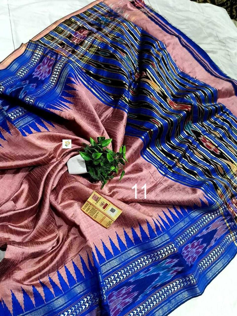 May include: A woven textile featuring a dusty rose center with a prominent blue border. The border displays intricate ikat patterns in black, white, and blue, accented by a sharp triangular motif. A small white tag with the number 11 is visible.