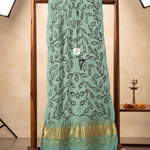 May include: A light mint green fabric drape featuring a repeating black embroidered leaf pattern and a wide metallic gold border at the bottom. The fabric is displayed on a wooden stand and includes a small white square label with the number 7.