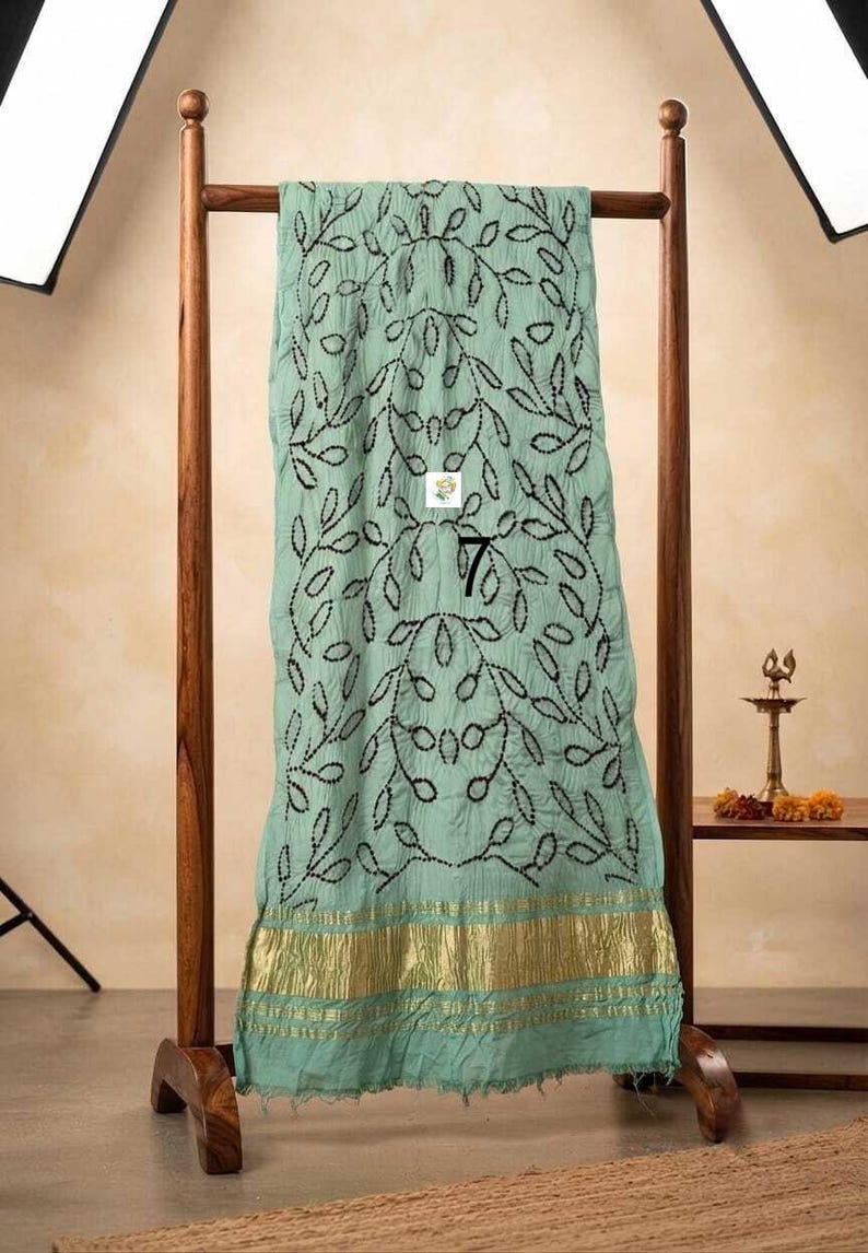 May include: A light mint green fabric drape featuring a repeating black embroidered leaf pattern and a wide metallic gold border at the bottom. The fabric is displayed on a wooden stand and includes a small white square label with the number 7.