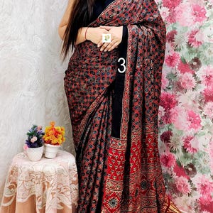 May include: A black saree featuring a dense, repeating geometric pattern in red, blue, and white. The fabric includes a wide black border and a pleated gold metallic hem. The number 3 is printed in white on the fabric, and a small white tag is visible.