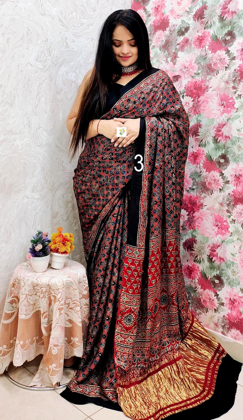 May include: A black saree featuring a dense, repeating geometric pattern in red, blue, and white. The fabric includes a wide black border and a pleated gold metallic hem. The number 3 is printed in white on the fabric, and a small white tag is visible.