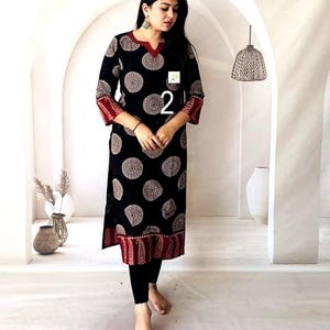 Puede incluir: Vestido t&uacute;nica negro con un estampado circular en blanco y beige. El vestido tiene ribetes rojos en el escote, las mangas y el dobladillo. El n&uacute;mero "2" es visible en la parte delantera. La modelo lleva leggings negros.