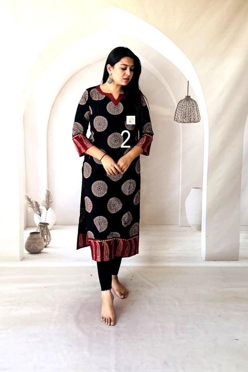 Puede incluir: Vestido t&uacute;nica negro con un estampado circular en blanco y beige. El vestido tiene ribetes rojos en el escote, las mangas y el dobladillo. El n&uacute;mero "2" es visible en la parte delantera. La modelo lleva leggings negros.