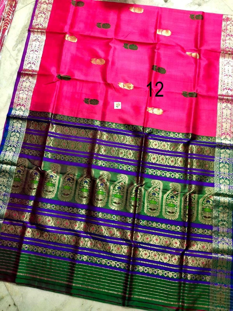 May include: A vibrant pink saree featuring small gold and green motifs scattered across the fabric. The wide border displays intricate purple and green bands with detailed gold zari paisley patterns and floral designs, finished with a silver edge.