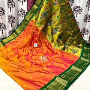 Op de afbeelding: Een levendige oranje en gele sari met een brede, donkergroene rand met gouden metalen details. De stof is versierd met kleine gele bloemmotieven en een groot, ingewikkeld goudkleurig paneel met kleurrijke pauwenpatronen en het getal 16.