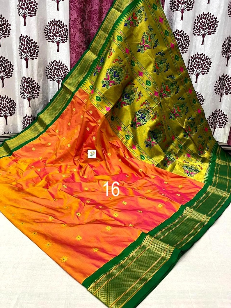 Op de afbeelding: Een levendige oranje en gele sari met een brede, donkergroene rand met gouden metalen details. De stof is versierd met kleine gele bloemmotieven en een groot, ingewikkeld goudkleurig paneel met kleurrijke pauwenpatronen en het getal 16.