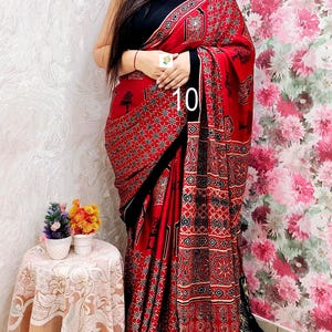 May include: A red saree featuring a black and white geometric and floral block print pattern, paired with a solid black blouse. The garment includes a black border and a metallic gold fringe trim at the end, with the number 10 printed on the fabric.