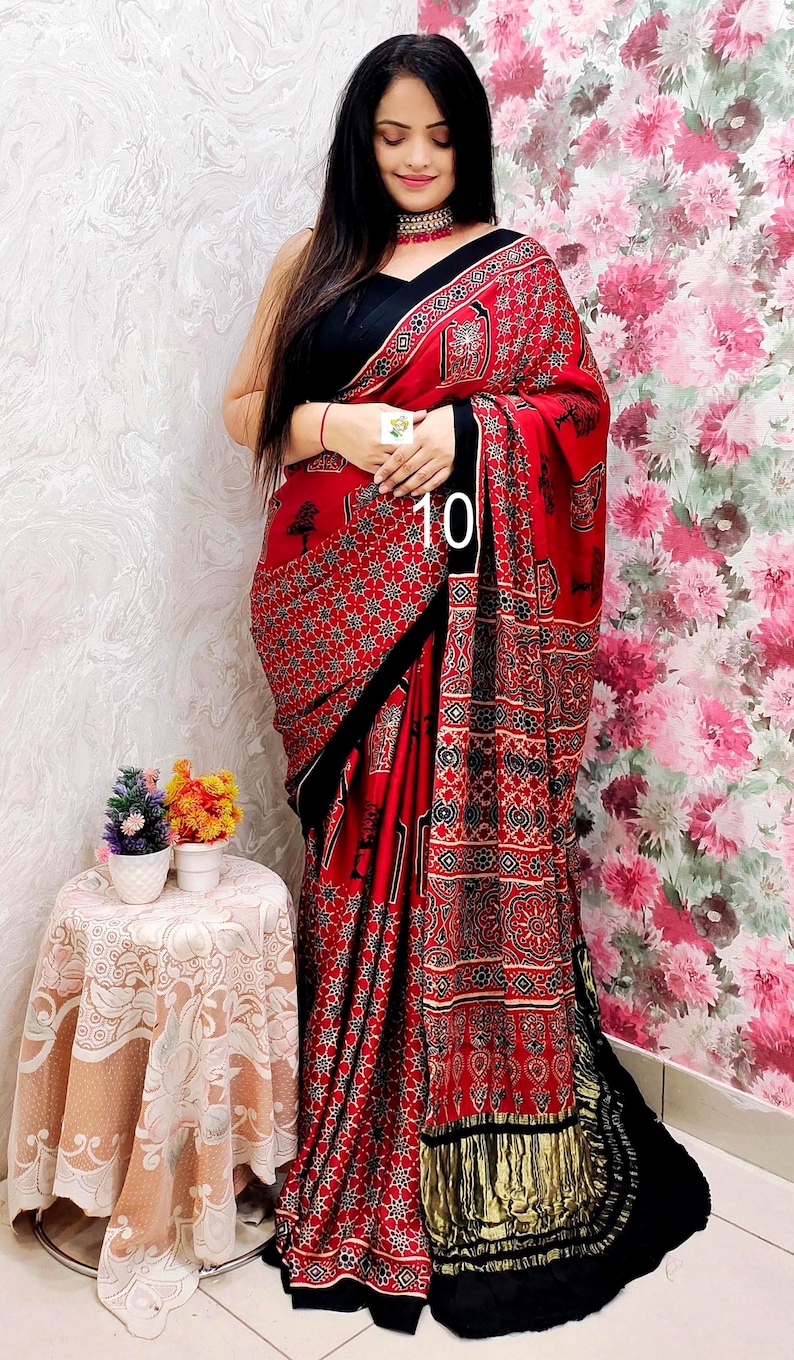 May include: A red saree featuring a black and white geometric and floral block print pattern, paired with a solid black blouse. The garment includes a black border and a metallic gold fringe trim at the end, with the number 10 printed on the fabric.