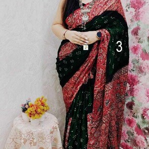 May include: A saree featuring a dark green section with small white dot patterns and a contrasting red border with intricate floral and geometric prints. The garment is draped over the body and includes a silver-toned necklace with a large pendant.