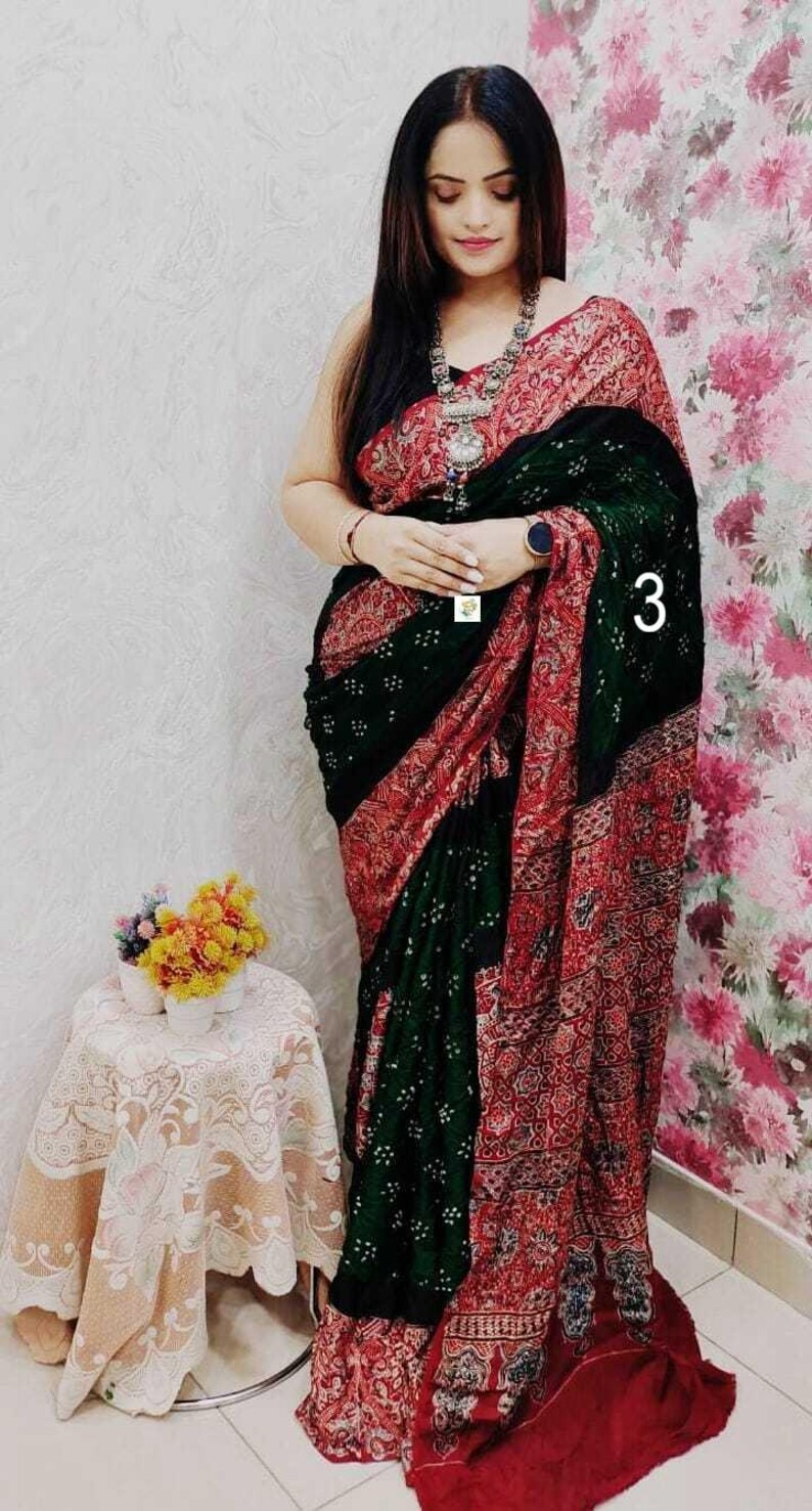 May include: A saree featuring a dark green section with small white dot patterns and a contrasting red border with intricate floral and geometric prints. The garment is draped over the body and includes a silver-toned necklace with a large pendant.