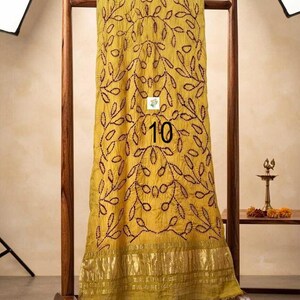 May include: A mustard yellow scarf featuring a dark brown embroidered leaf pattern and a metallic gold border at the bottom. The fabric has a crinkled texture and displays the number 10 printed in black on the center of the fabric surface.