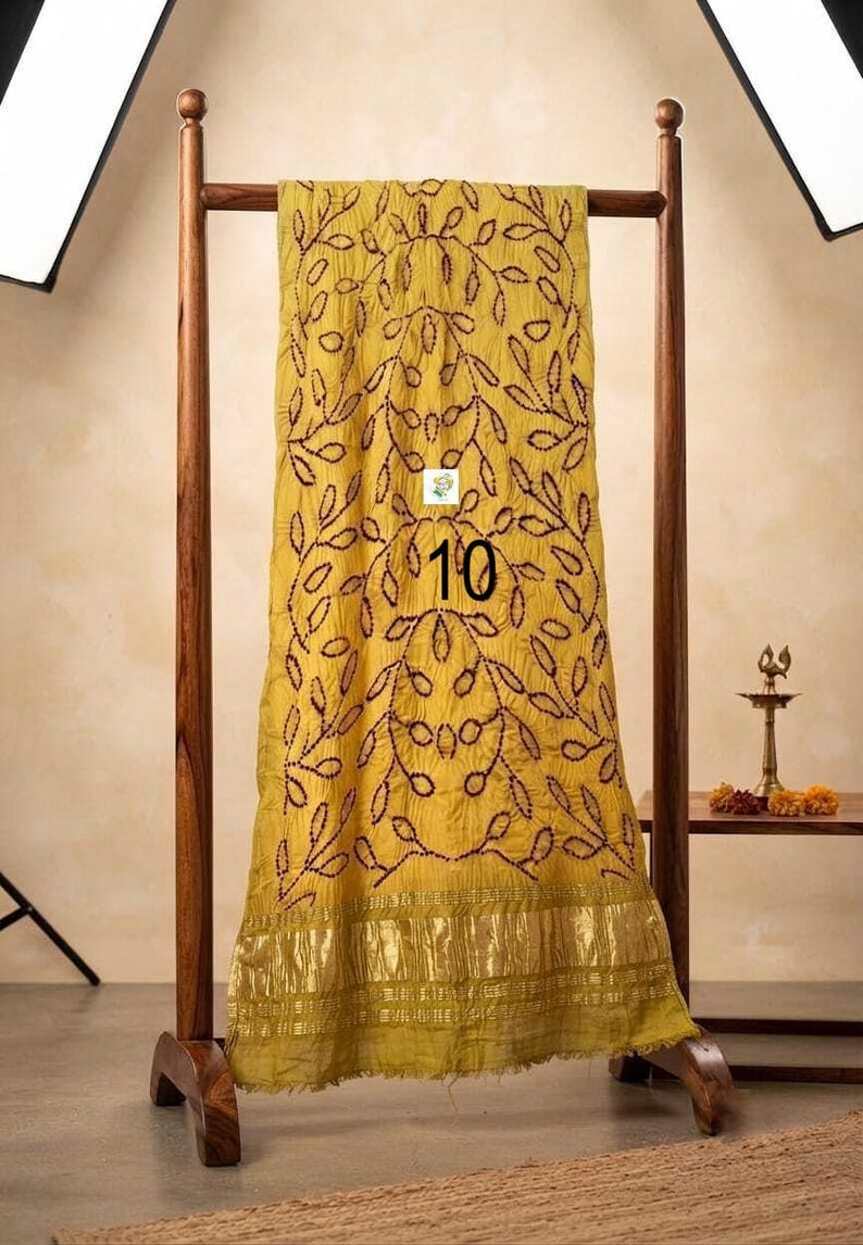 May include: A mustard yellow scarf featuring a dark brown embroidered leaf pattern and a metallic gold border at the bottom. The fabric has a crinkled texture and displays the number 10 printed in black on the center of the fabric surface.