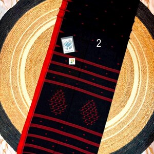 May include: A black textile featuring a bold red border and horizontal red stripes. The fabric is decorated with small red dots and two prominent red diamond-shaped patterns. A small square label with the number 2 is visible on the surface.