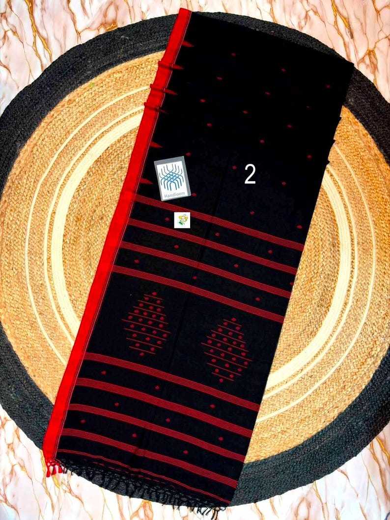 May include: A black textile featuring a bold red border and horizontal red stripes. The fabric is decorated with small red dots and two prominent red diamond-shaped patterns. A small square label with the number 2 is visible on the surface.