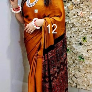 Puede incluir: Un sari de color amarillo mostaza con un borde estampado en marr&oacute;n oscuro y rojo con intrincados dise&ntilde;os geom&eacute;tricos. La prenda est&aacute; drapeada en un estilo tradicional, complementada con un collar de cuentas blancas y pulseras a juego.