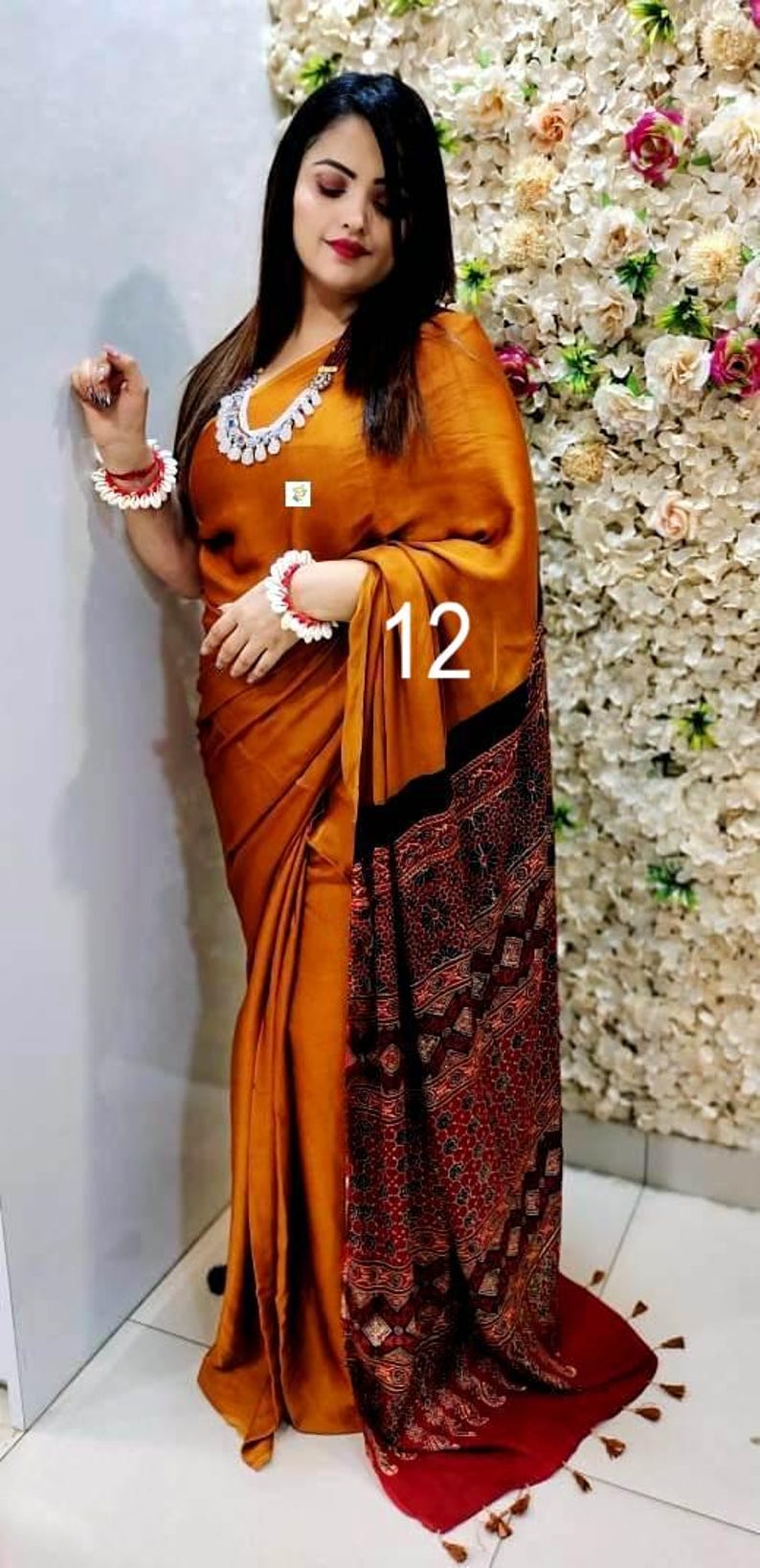 Puede incluir: Un sari de color amarillo mostaza con un borde estampado en marr&oacute;n oscuro y rojo con intrincados dise&ntilde;os geom&eacute;tricos. La prenda est&aacute; drapeada en un estilo tradicional, complementada con un collar de cuentas blancas y pulseras a juego.