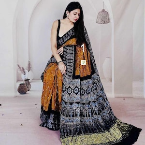 May include: A saree featuring a mustard yellow base with a black border and pallu. The fabric displays intricate black and white geometric patterns, floral motifs, and tree designs. The garment includes a sleeveless black blouse with a scoop neckline.