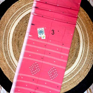 May include: A rectangular pink fabric textile featuring horizontal white stripes, small scattered white dots, and two diamond-shaped patterns made of white dots. A small white tag with a blue graphic and the number 3 are attached to the fabric.