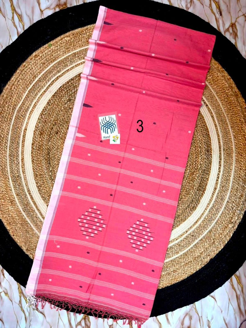 May include: A rectangular pink fabric textile featuring horizontal white stripes, small scattered white dots, and two diamond-shaped patterns made of white dots. A small white tag with a blue graphic and the number 3 are attached to the fabric.