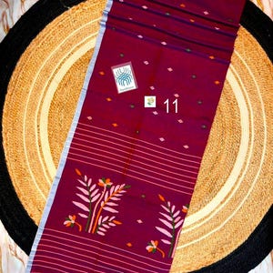 May include: A deep maroon textile featuring a light blue border, horizontal white stripes, and small diamond-shaped motifs. The fabric displays a large floral pattern with pink and orange leaves and includes a tag labeled Handloom and the number 11.