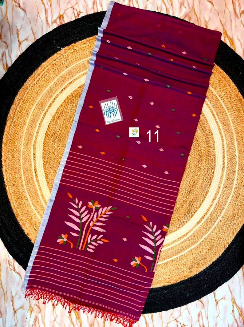 May include: A deep maroon textile featuring a light blue border, horizontal white stripes, and small diamond-shaped motifs. The fabric displays a large floral pattern with pink and orange leaves and includes a tag labeled Handloom and the number 11.
