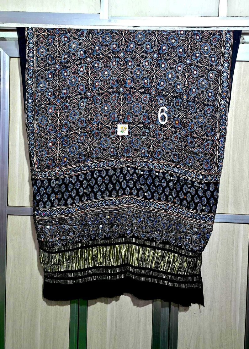 Może przedstawiać: Czarna tkanina z misternym wzorem niebieskich i złotych motyw&oacute;w paisley, kształt&oacute;w geometrycznych oraz małych, odblaskowych akcent&oacute;w lustrzanych. Dolna część posiada wyraźny poziomy pas o złotej, plisowanej fakturze.