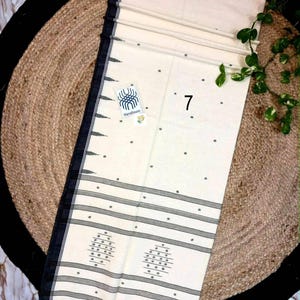 May include: An off-white handloom saree featuring a black border with geometric patterns, small dark grey polka dots across the fabric, and horizontal black stripes near the hem. A small tag with the number 7 and a handloom logo is attached.