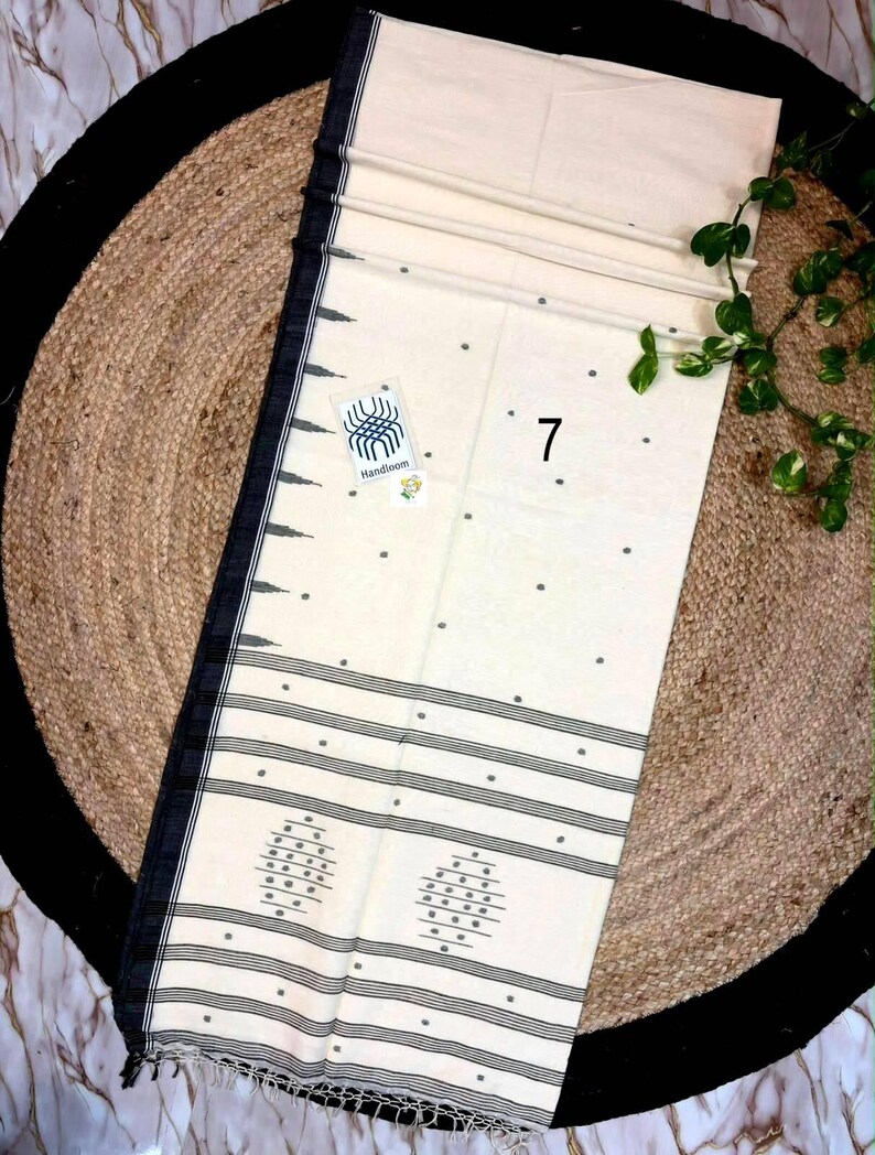 May include: An off-white handloom saree featuring a black border with geometric patterns, small dark grey polka dots across the fabric, and horizontal black stripes near the hem. A small tag with the number 7 and a handloom logo is attached.