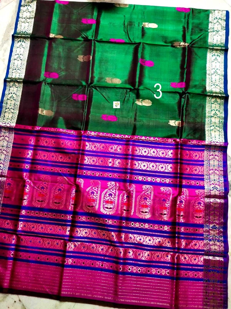 May include: A vibrant green and magenta silk saree featuring a wide, ornate border with intricate gold zari patterns, paisley motifs, and horizontal stripes. The main body displays scattered gold and magenta floral accents and the number 3.