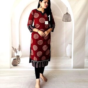 Puede incluir: Un vestido t&uacute;nica burdeos con un estampado circular crema. El vestido tiene mangas tres cuartos con un estampado de cachemira negro y crema en los pu&ntilde;os. El escote y el dobladillo tambi&eacute;n presentan detalles en negro y crema. La modelo lleva leggings negros.