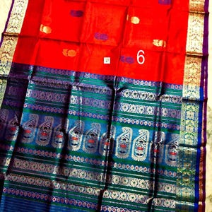 May include: A vibrant red saree featuring a wide border with intricate blue, green, and metallic gold woven patterns. The fabric displays small scattered motifs in gold and purple, with a prominent white square label displaying the number 6.