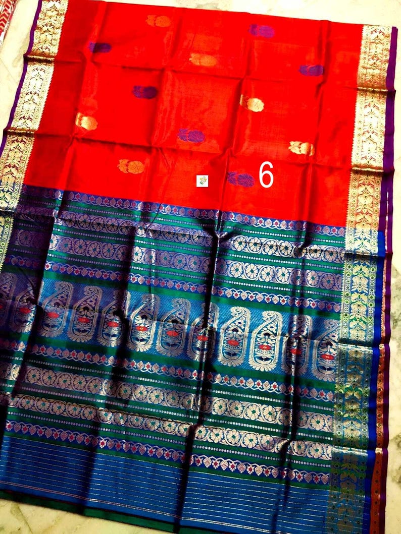 May include: A vibrant red saree featuring a wide border with intricate blue, green, and metallic gold woven patterns. The fabric displays small scattered motifs in gold and purple, with a prominent white square label displaying the number 6.