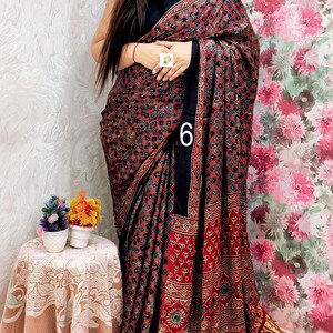 May include: A black saree featuring a dense, repeating geometric pattern in red, blue, and white. The garment includes a solid black border and a metallic gold pleated hem. The number 6 is printed on the fabric, and the design includes intricate motifs.