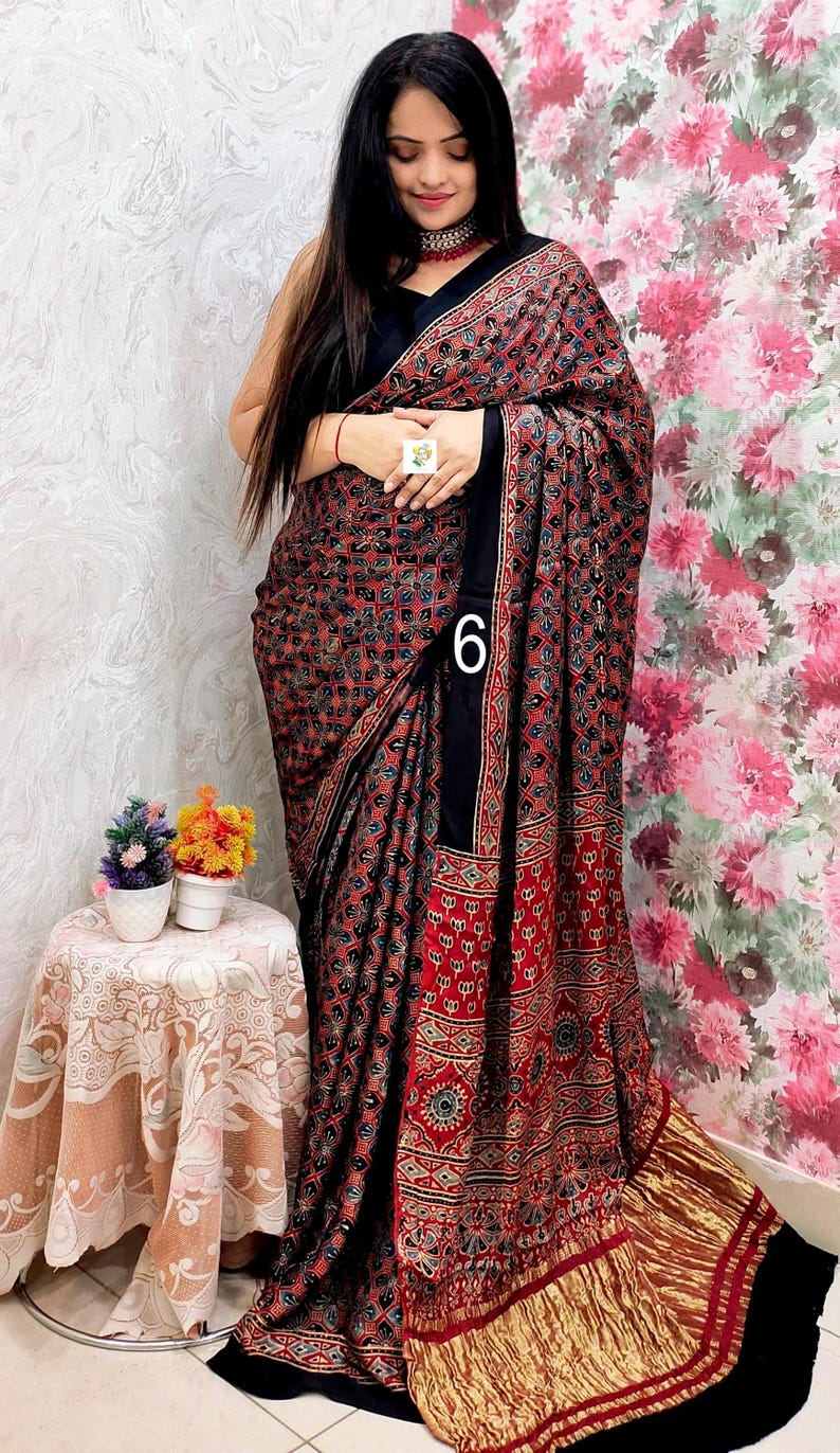 May include: A black saree featuring a dense, repeating geometric pattern in red, blue, and white. The garment includes a solid black border and a metallic gold pleated hem. The number 6 is printed on the fabric, and the design includes intricate motifs.