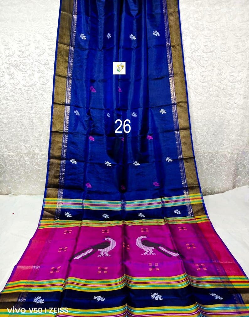 Puede incluir: Un sari azul real con un borde ancho magenta que presenta dos motivos de pavo real en blanco y negro. La tela muestra peque&ntilde;os patrones florales blancos en todo el cuerpo, rematado con ribetes met&aacute;licos dorados y rayas horizontales amarillas y verdes.