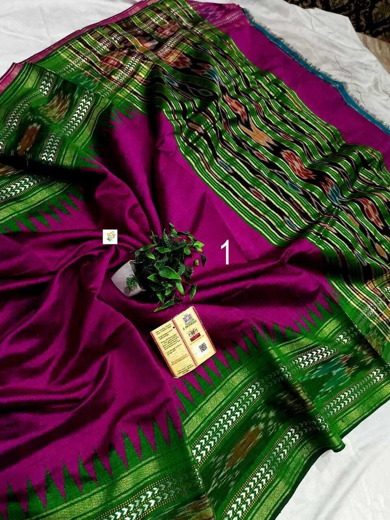 May include: A vibrant magenta saree featuring a wide, deep green border with intricate ikat patterns and a metallic gold zari edge. The fabric displays a smooth texture, accented by a small green plant and a rectangular product tag labeled with the number 1.