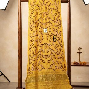 May include: A bright yellow rectangular scarf featuring a dark brown stitched leaf pattern throughout the fabric. The scarf includes a wide metallic gold border at the bottom edge and displays the number 6 printed in black on the center surface.