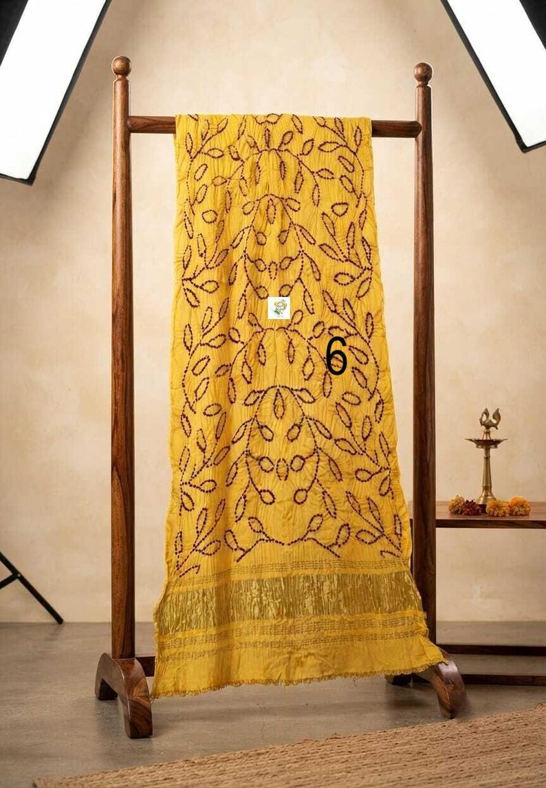 May include: A bright yellow rectangular scarf featuring a dark brown stitched leaf pattern throughout the fabric. The scarf includes a wide metallic gold border at the bottom edge and displays the number 6 printed in black on the center surface.
