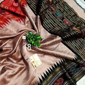 May include: A beige saree featuring a black border with intricate ikat patterns in red, green, and yellow. The fabric has a subtle sheen, and the border includes a sharp triangular motif. A small tag with the number 18 is visible on the fabric.