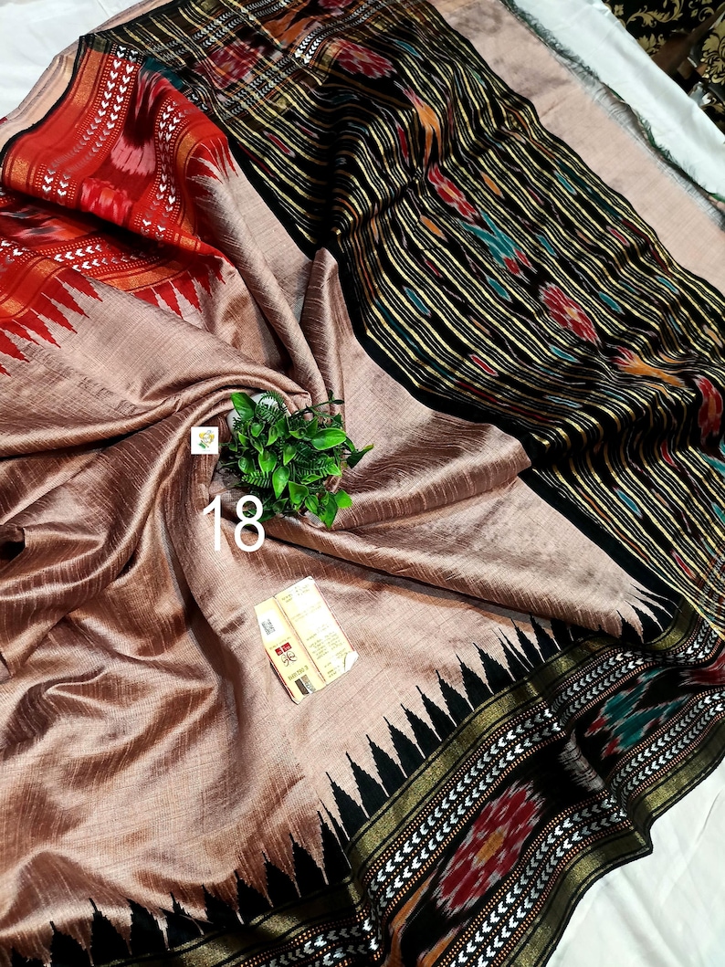 May include: A beige saree featuring a black border with intricate ikat patterns in red, green, and yellow. The fabric has a subtle sheen, and the border includes a sharp triangular motif. A small tag with the number 18 is visible on the fabric.