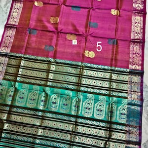 May include: A magenta saree featuring gold floral motifs and a large teal border with intricate gold paisley patterns and horizontal stripes. The fabric displays a woven texture with a prominent number 5 printed on the magenta section.