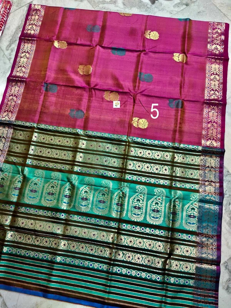 May include: A magenta saree featuring gold floral motifs and a large teal border with intricate gold paisley patterns and horizontal stripes. The fabric displays a woven texture with a prominent number 5 printed on the magenta section.