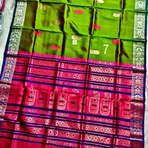 May include: A vibrant green silk saree featuring a wide, ornate border in hot pink and deep blue. The fabric is decorated with small gold-toned floral motifs and a prominent white tag with the number 7 printed on the center of the green section.