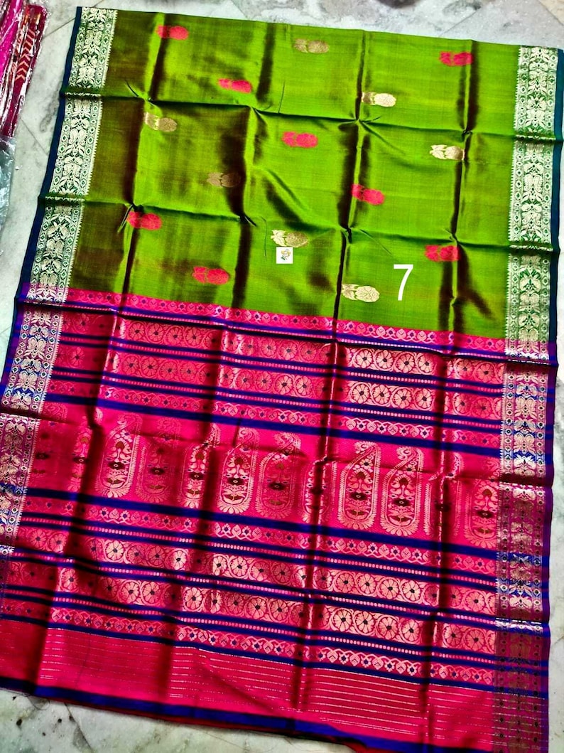 May include: A vibrant green silk saree featuring a wide, ornate border in hot pink and deep blue. The fabric is decorated with small gold-toned floral motifs and a prominent white tag with the number 7 printed on the center of the green section.