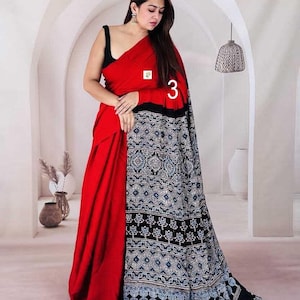 Puede incluir: Un sari rojo vibrante con un cuerpo rojo s&oacute;lido y un extremo negro contrastante adornado con intrincados patrones geom&eacute;tricos y florales en blanco y azul. La prenda incluye una peque&ntilde;a etiqueta cuadrada blanca y el n&uacute;mero 3 en blanco.