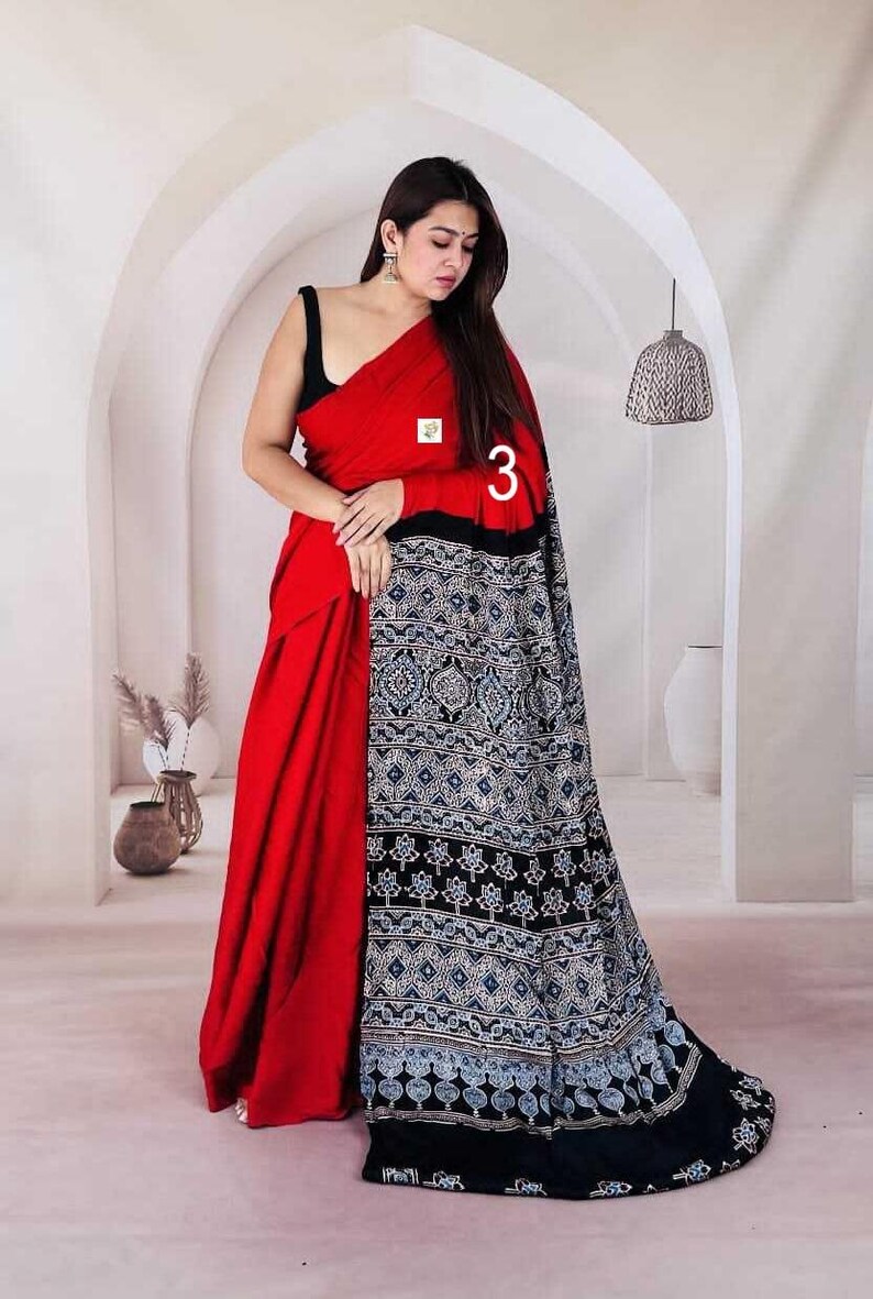 Puede incluir: Un sari rojo vibrante con un cuerpo rojo s&oacute;lido y un extremo negro contrastante adornado con intrincados patrones geom&eacute;tricos y florales en blanco y azul. La prenda incluye una peque&ntilde;a etiqueta cuadrada blanca y el n&uacute;mero 3 en blanco.