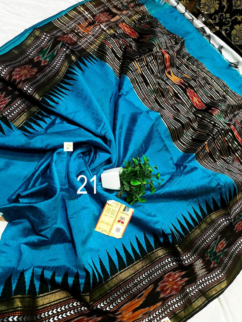 May include: A bright blue saree featuring a wide black border with intricate ikat patterns in red, white, and green. The fabric has a subtle sheen and a prominent black triangular motif along the edge. A small white tag with the number 21 is visible.