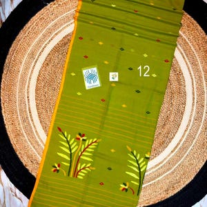 May include: An olive green textile featuring a yellow border, small diamond-shaped patterns in red and yellow, and a large floral motif at the bottom. The fabric includes thin horizontal stripes, a Handloom label, and the number 12 printed on it.