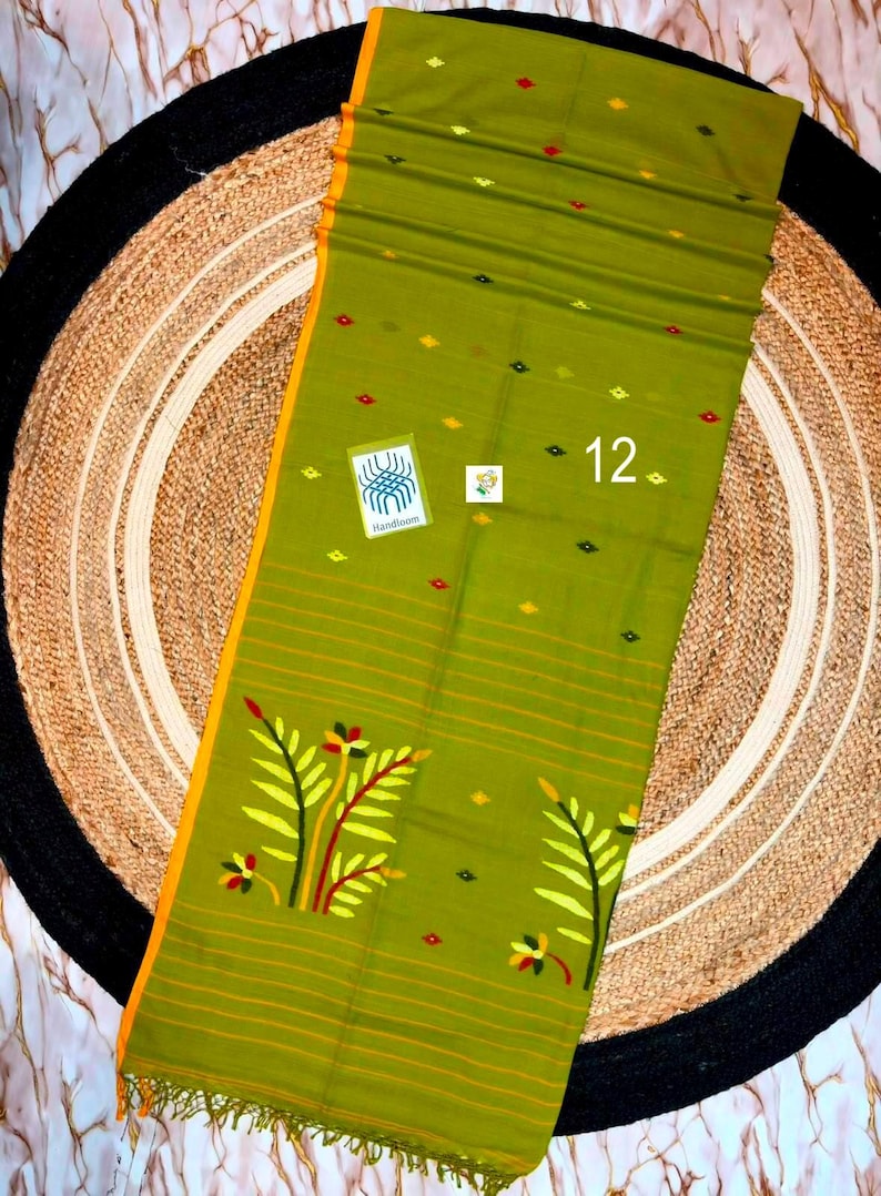 May include: An olive green textile featuring a yellow border, small diamond-shaped patterns in red and yellow, and a large floral motif at the bottom. The fabric includes thin horizontal stripes, a Handloom label, and the number 12 printed on it.